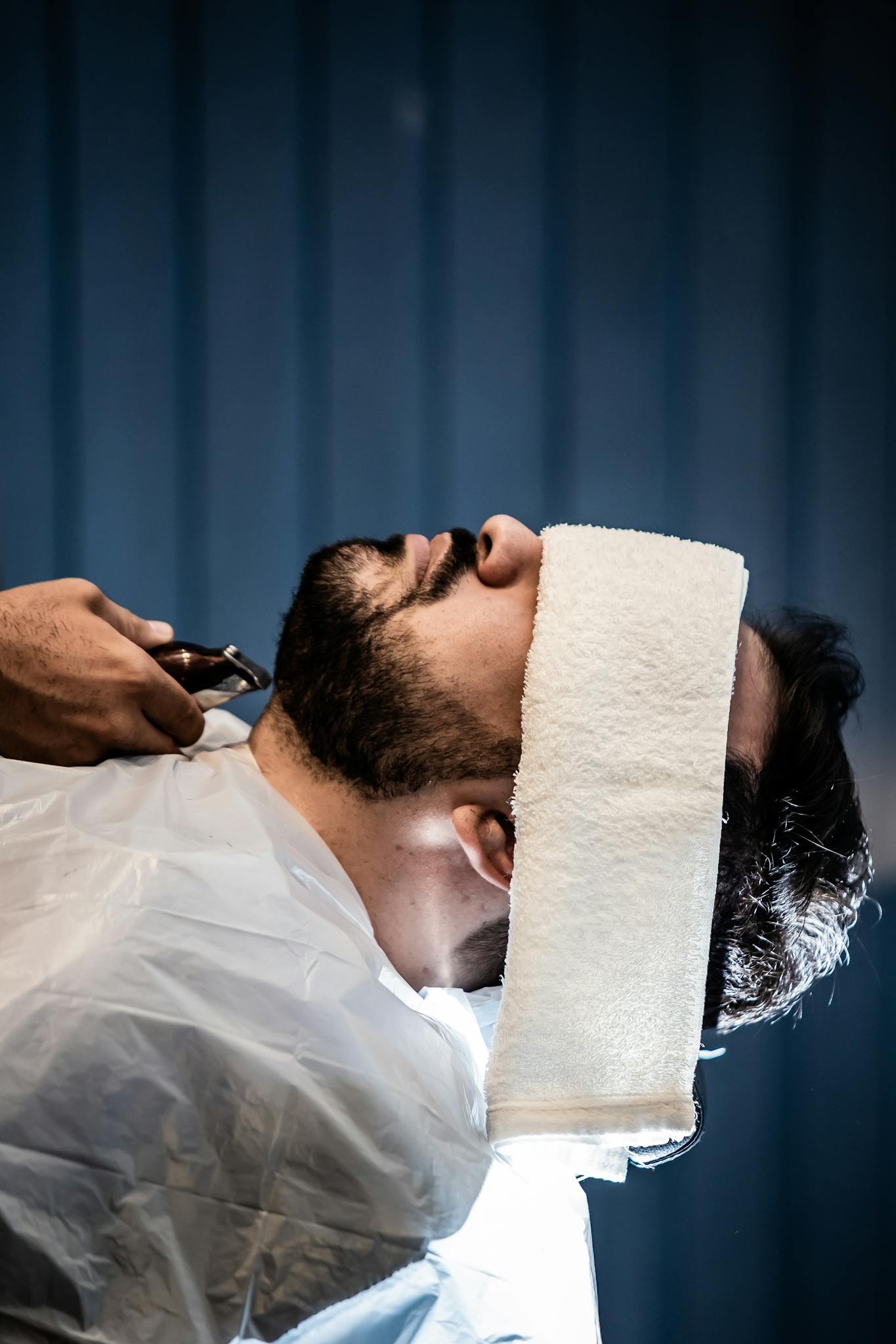 A professional barber shaving and styling a bearded man's face indoors at a salon.